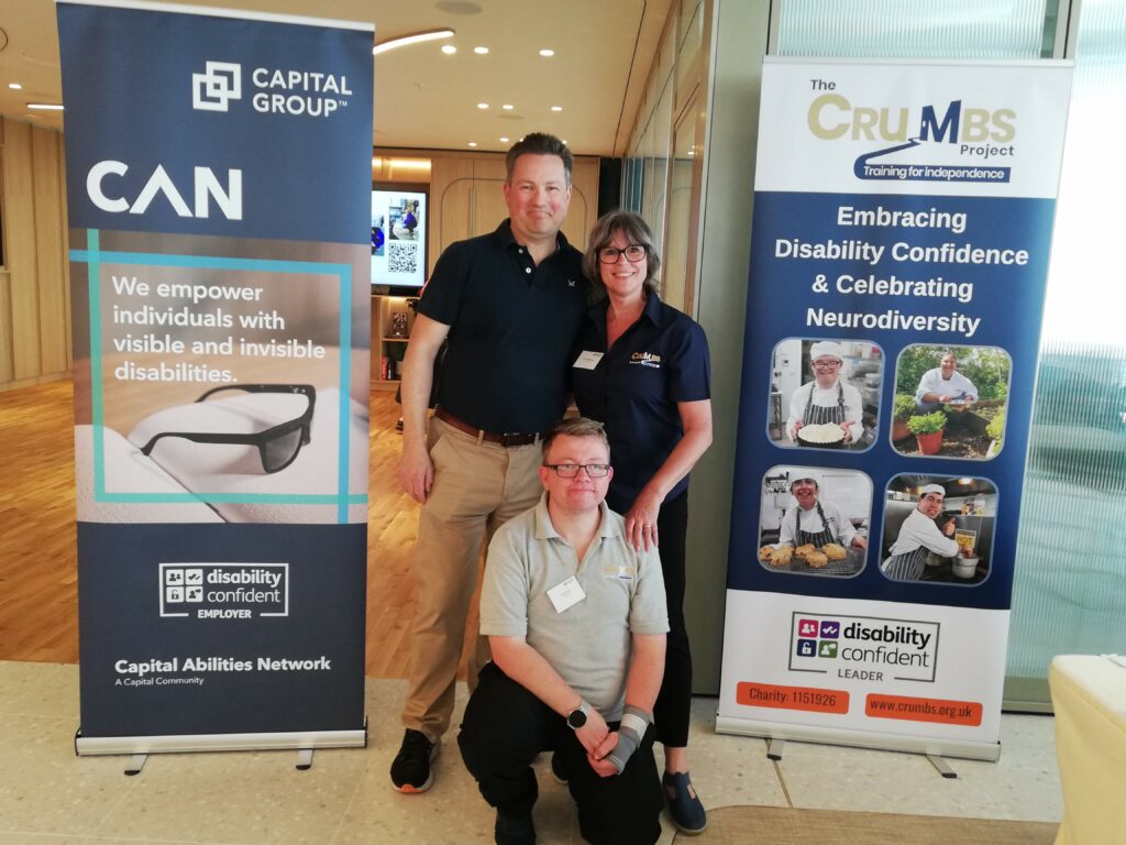 Russell from Capital Group stands next to Ursula from The CRUMBS Project. Former trainee Jake knees in front of them between two marketing banners.