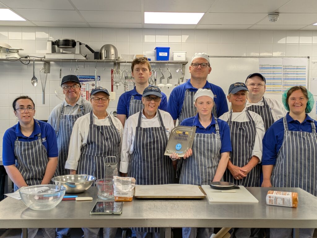 Trainees and staff pose with a Craft Guild of Chefs' plaque in the training bakery at The CRUMBS Project