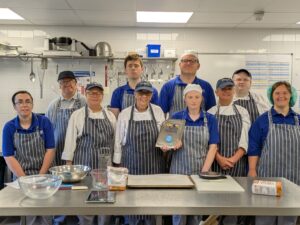 Trainees and staff pose with a Craft Guild of Chefs' plaque in the training bakery at The CRUMBS Project